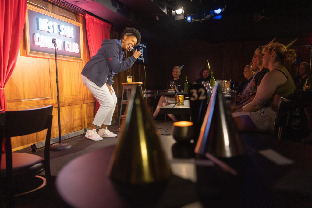 Comic Joyelle Nicole Johnson on stage at a comedy show in front of an audience wearing party hats