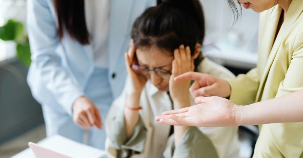 woman with glasses sits at a desk looking frustrated with her hands on her face with two women next to her out of frame