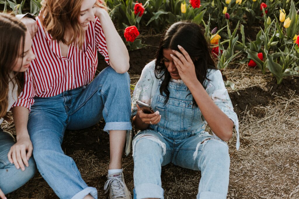 Three young women sit together on the ground in a garden; one looks at a smartphone while the others watch, with tulips in the background.