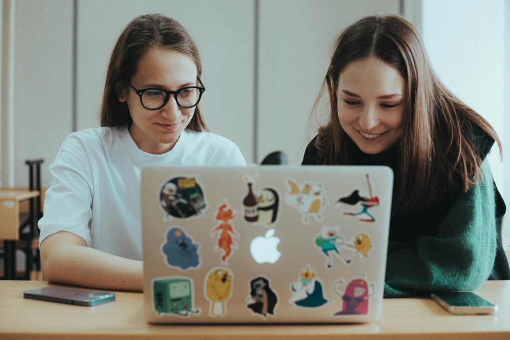Two women viewing a screen on a sticker-decorated laptop.
