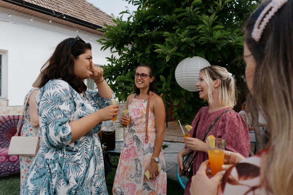 Four women are gathered outdoors, smiling and holding glasses with orange drinks. A white paper lantern hangs in the background.
