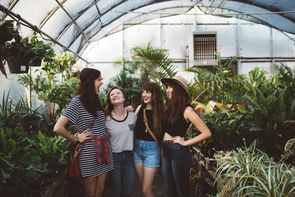 A group of four women smiling and talking closely together among rows of plants in a bright greenhouse.