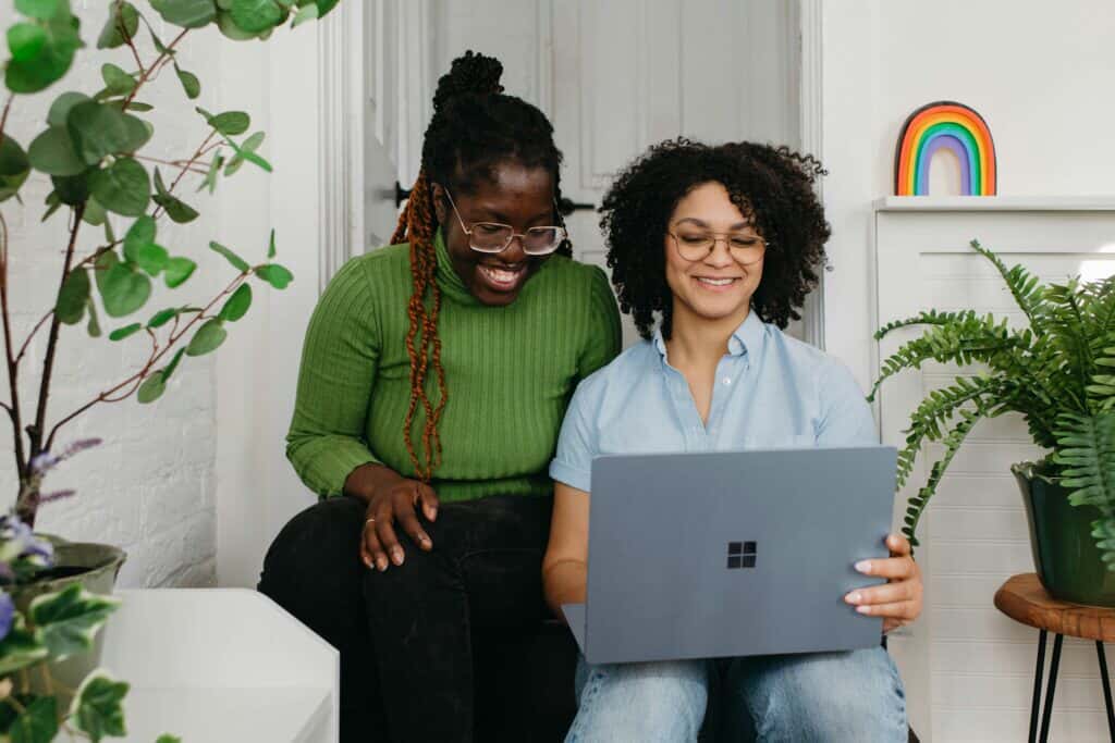 Two women smiling while working together on a laptop in a cozy indoor space decorated with plants and a rainbow ornament.