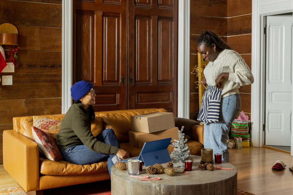 Two women share a joyful moment in a cozy, wood-paneled living room during the holidays. One woman, seated on a tan leather couch, smiles while the other stands holding a striped gift bag. Holiday decorations, a small silver Christmas tree, and gift boxes surround them.q