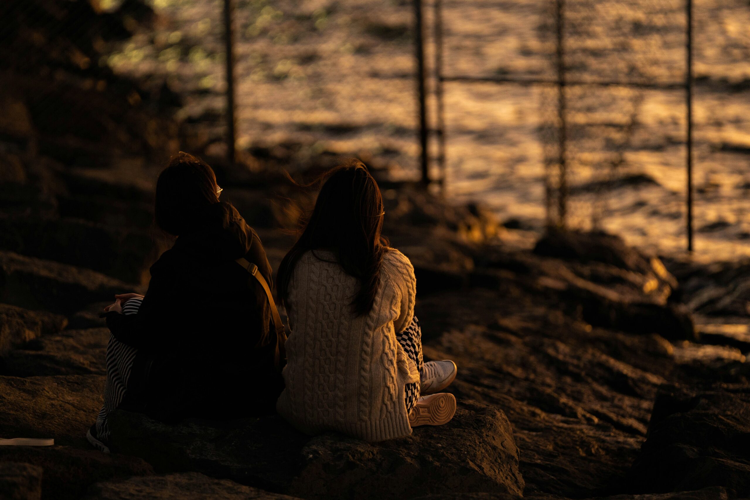 Two people sit quietly on rocks by the water during sunset, facing the glowing horizon. Their backs are turned, creating a reflective and peaceful mood.