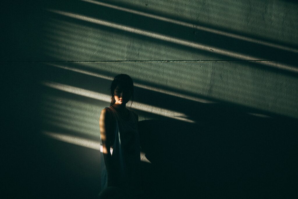 A person stands partially in shadow against a wall, with dramatic light streaming through blinds creating diagonal patterns. The mood is introspective and somber.