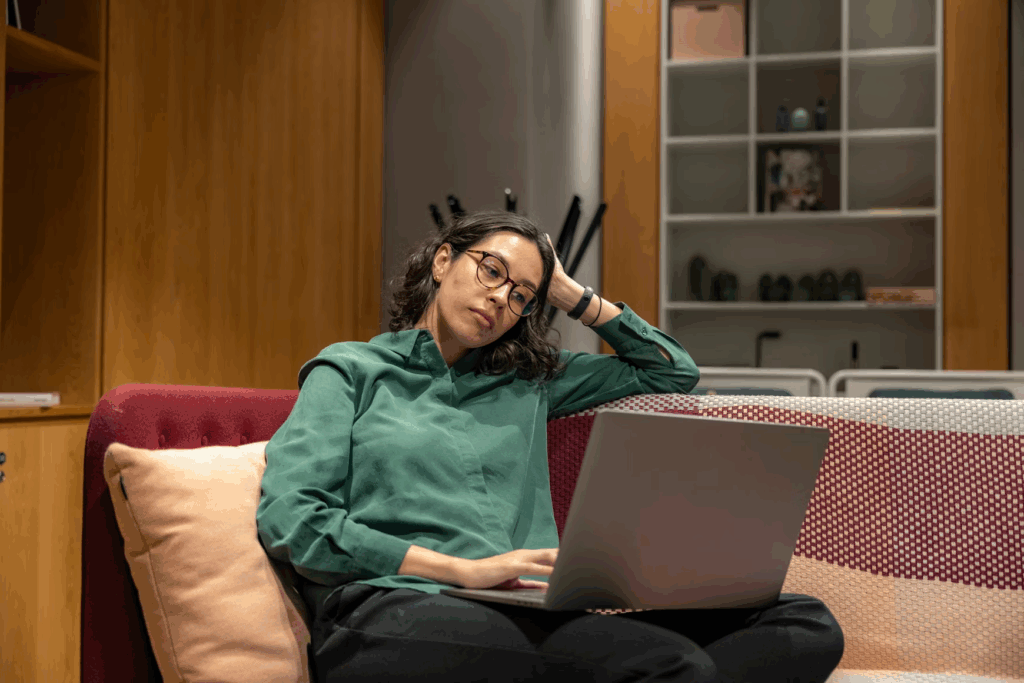 A woman sitting on a couch with a laptop on her lap, resting her head on her hand and appearing tired or stressed while working.