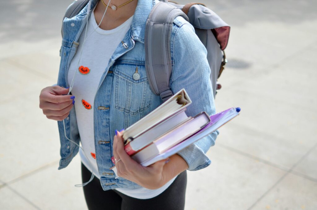 Student wearing a denim jacket and backpack holding books and notebooks while walking on campus.