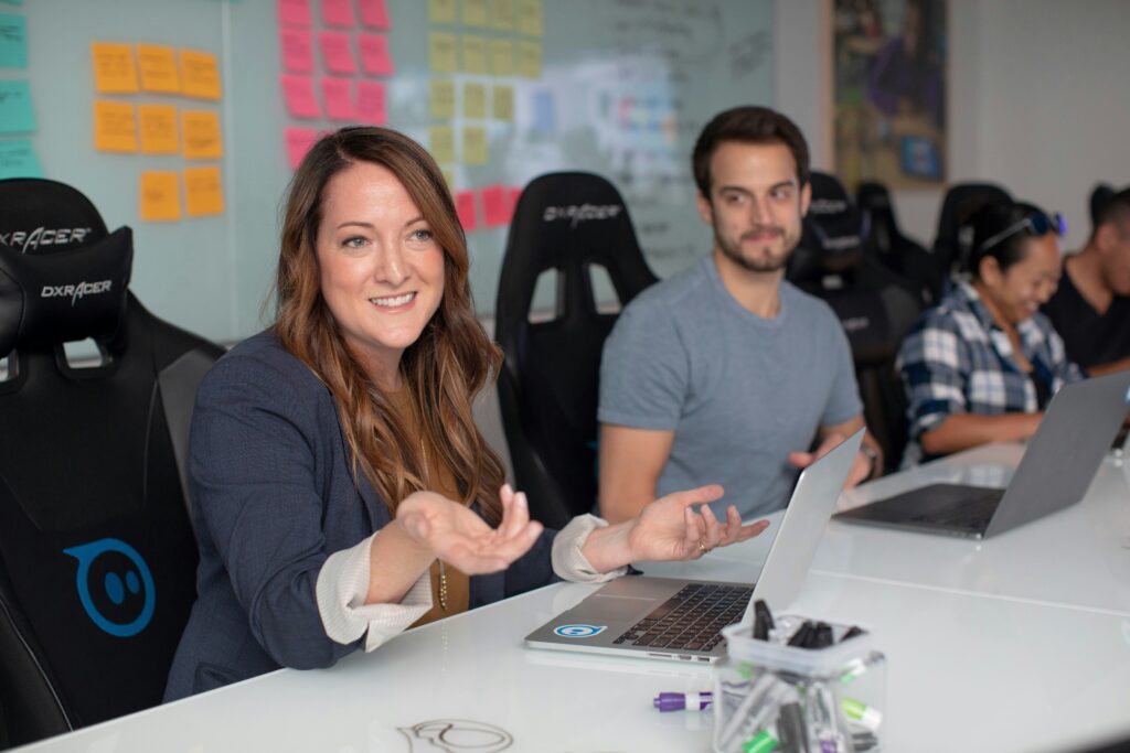 Colleagues collaborate in a busy, modern open-plan office, with two women smiling and talking at a desk in the foreground.