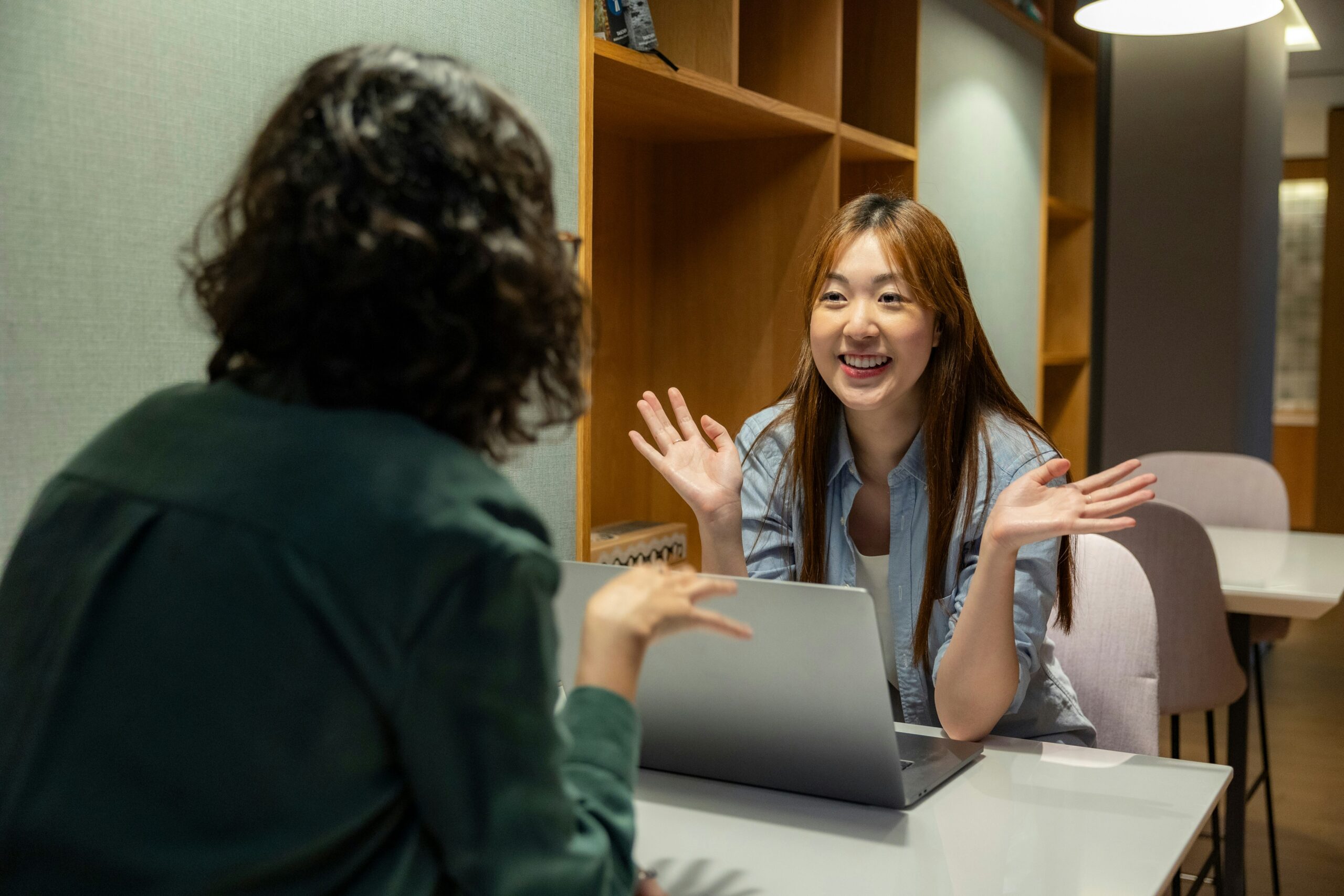 A young woman smiles and gestures enthusiastically during a positive job interview or collaborative meeting with a colleague across a table.