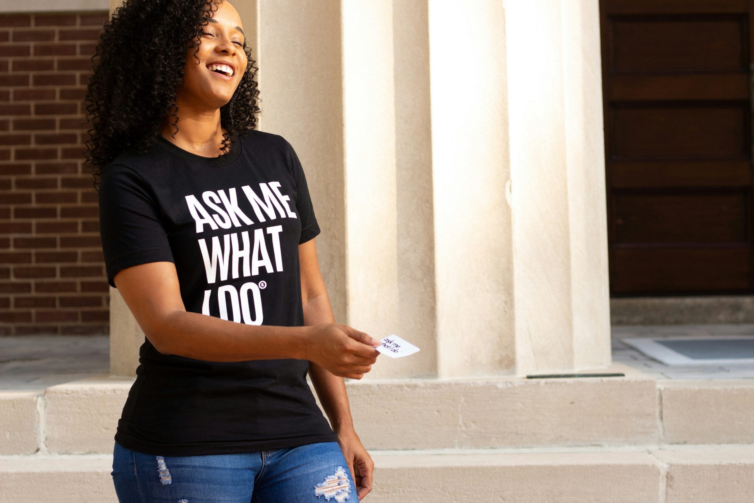 Woman with a shirt saying "Ask me what I do" and a business card she is handing out.